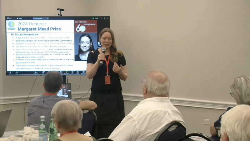 This photograph depicts the 2024 awards ceremony where Claudia Westermann is being honored with the Margaret Mead Prize of the American Society for Cybernetics, speaking to her colleagues and peers at the society's 60th anniversary meeting. Claudia Westermann in a black dress is standing at the front of the room, holding a microphone and speaking to an audience. Behind her is a large screen displaying a presentation slide about the "2024 Honoree Margaret Mead Prize." The slide includes some text and a black-and-white portrait photograph of herself. In the foreground, you can see the backs of several audience members seated at tables. The room itself is a meeting room with white walls and some decorative molding visible. On the tables in front of the audience, there are water bottles and conference materials.